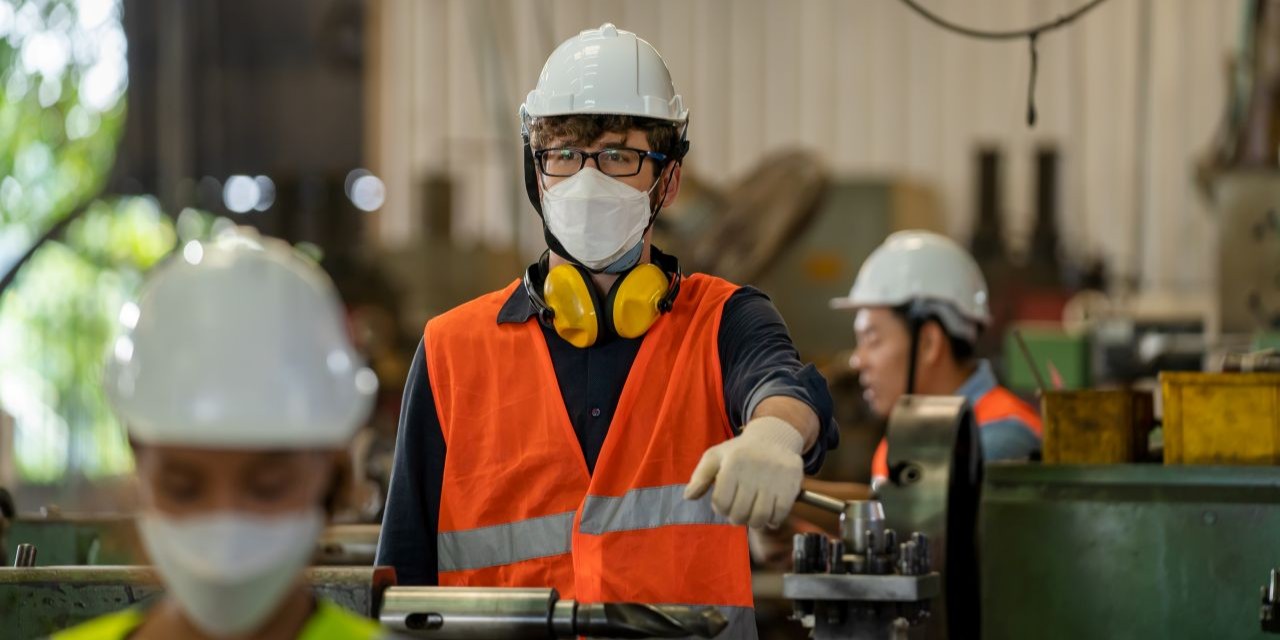 A wide industrial manufacturing plant interior showing workers in safety gear near production equipment, with visible dust collection/fume extraction hoods and ductwork overhead, clean air flow indicators or ventilation systems highlighted, soft natural or bright workshop lighting, modern and professional atmosphere (no faces needed if preferred for stock versatility).