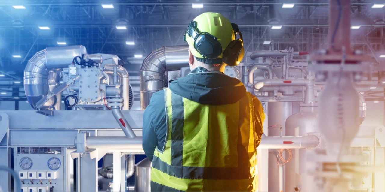 A factory or industrial workshop interior with workers wearing hearing protection near loud machinery, a sound level meter or noise dosimeter visible in the foreground or being used by a technician, bright industrial lighting, clear view of equipment and safety gear, professional and realistic workplace atmosphere.